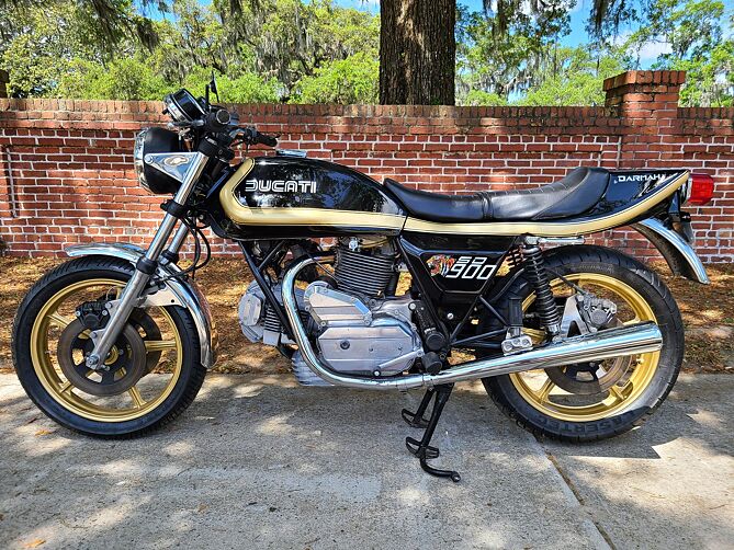 A vintage Ducati 900 motorcycle parked outdoors on a concrete surface in front of a brick wall and trees. The motorcycle features a black body with gold pinstripes, gold-colored wheels, and chrome engine components.